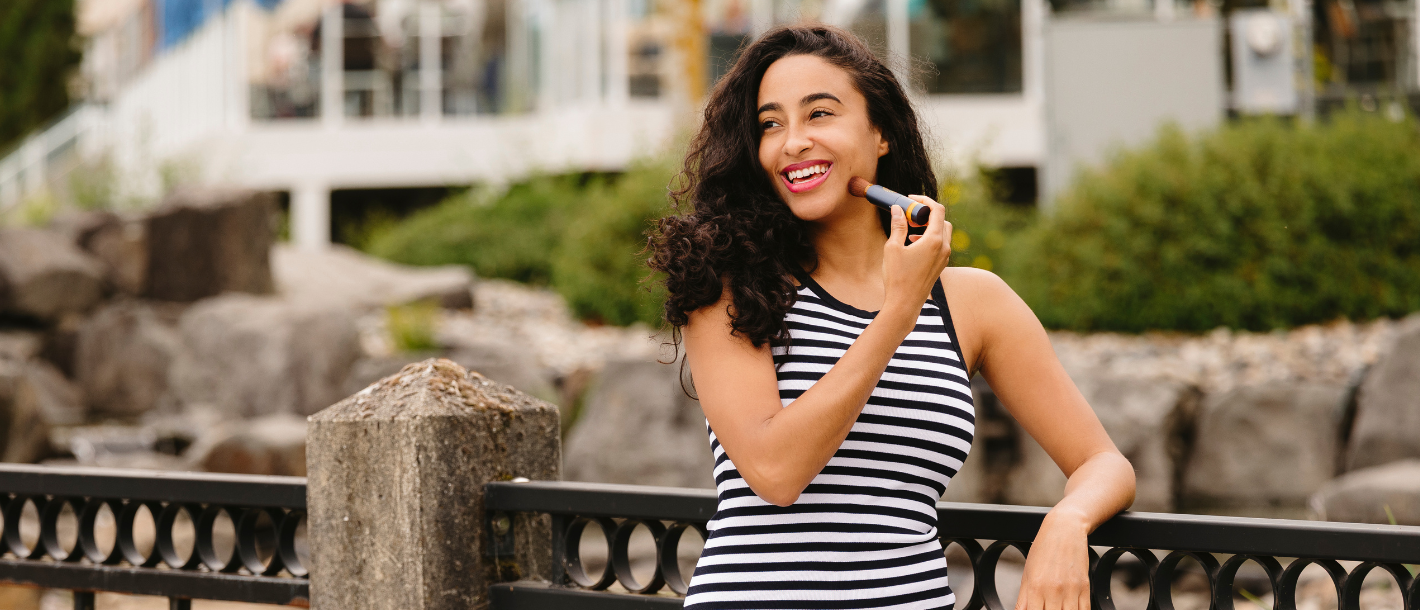 Woman outdoors smiling while applying Brush On Block® mineral powder sunscreen to her face during the day.
