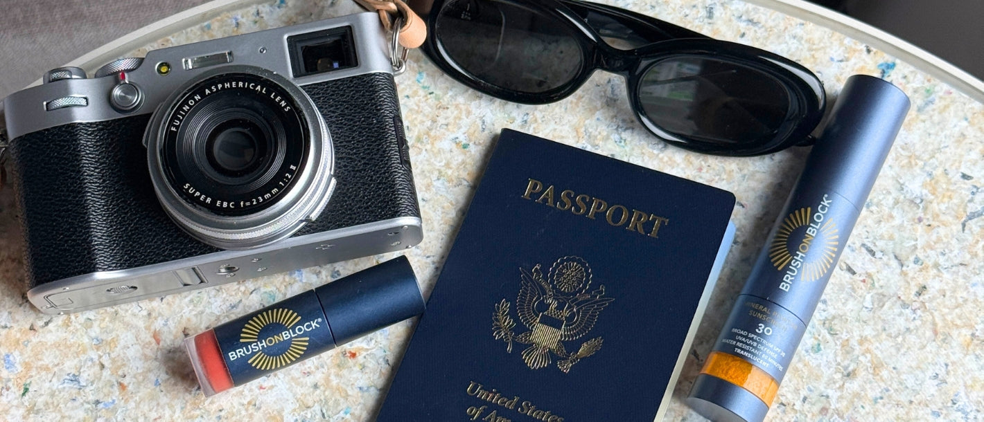 Brush On Block mineral powder, lip oil, passport, sunglasses, and camera on an airport table showing TSA friendly travel SPF essentials