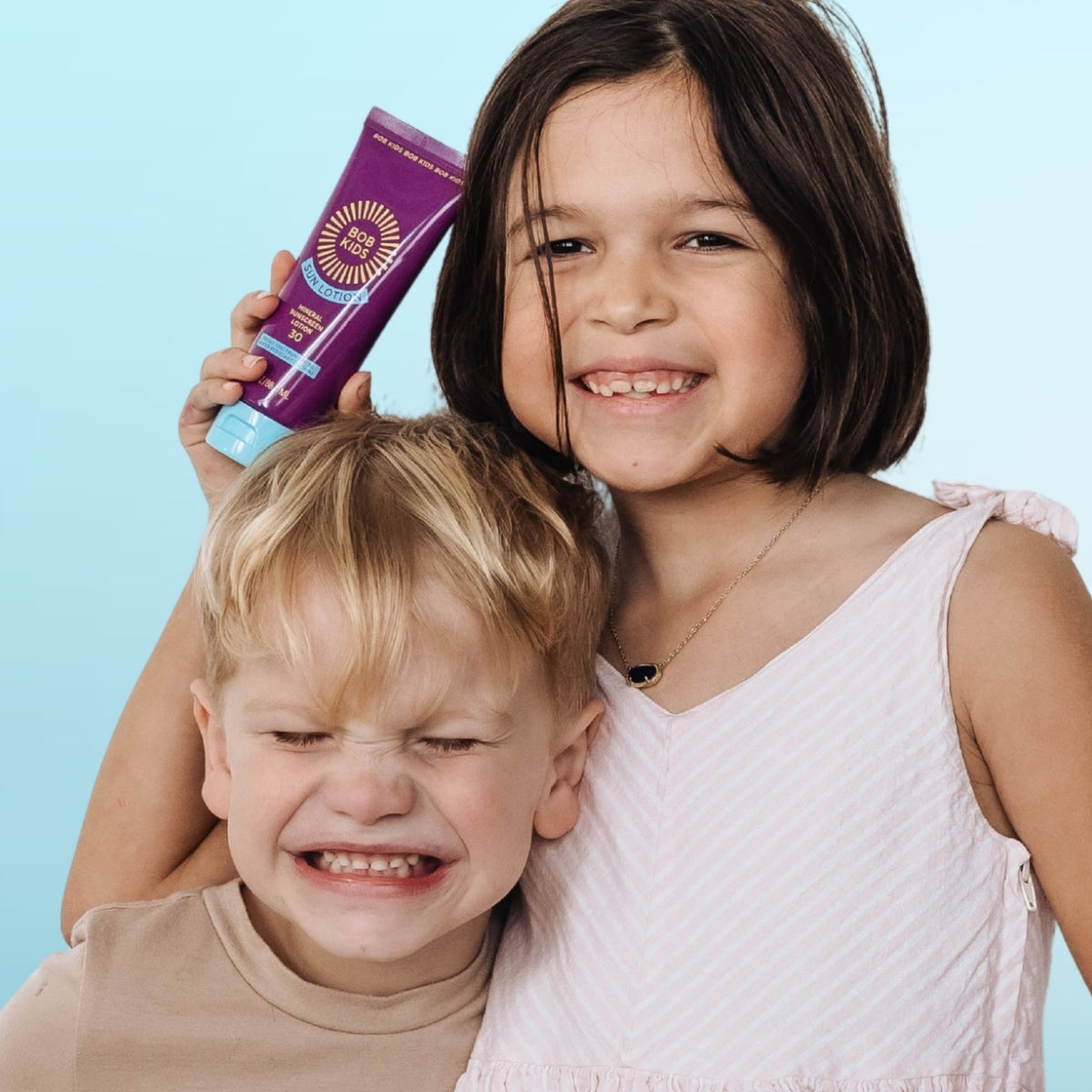 Two children with a purple bottle of BOB KIDS SPF 30 Mineral Sun Lotion.