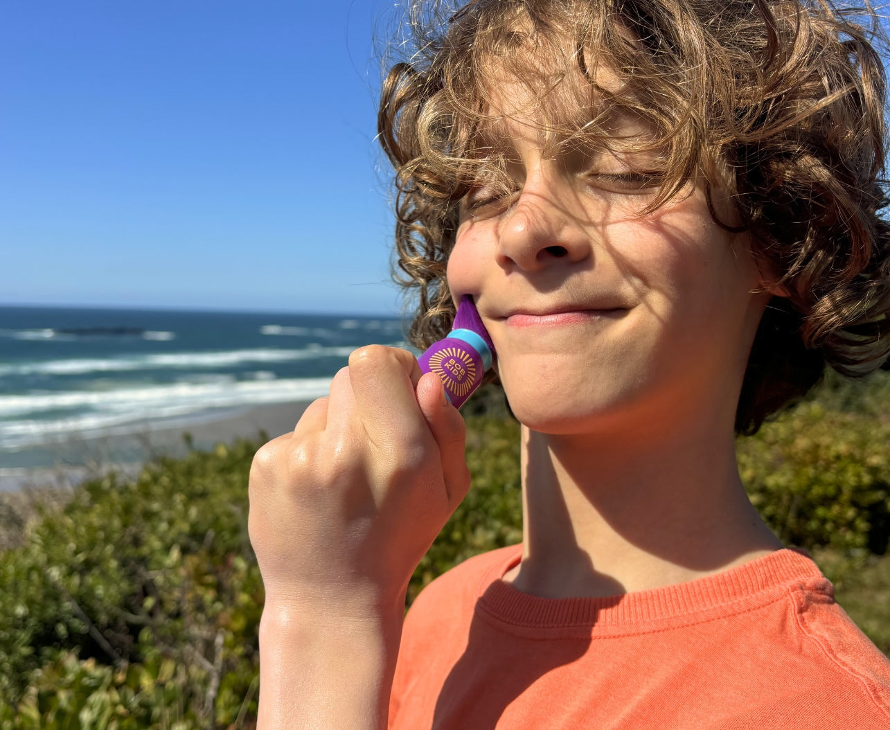 Boy applying BOB KIDS mineral powder sunscreen to his cheek at the beach