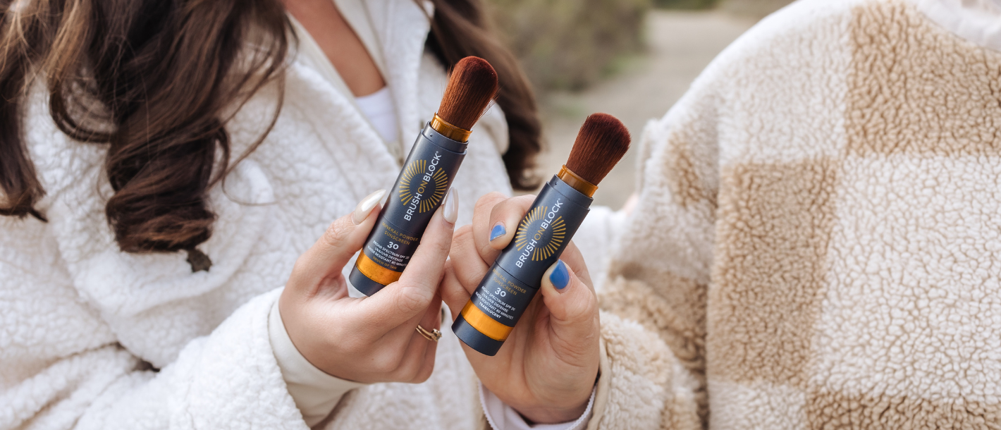 Two women outside holding up sunscreen brushes representing the everyday moment of remembering to reapply sunscreen