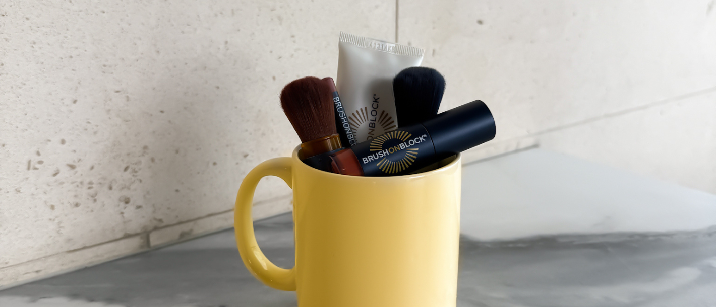 A coffee cup holding Brush On Block sunscreen essentials including a powder brush lip oil and lotion for a simple daily sun protection routine