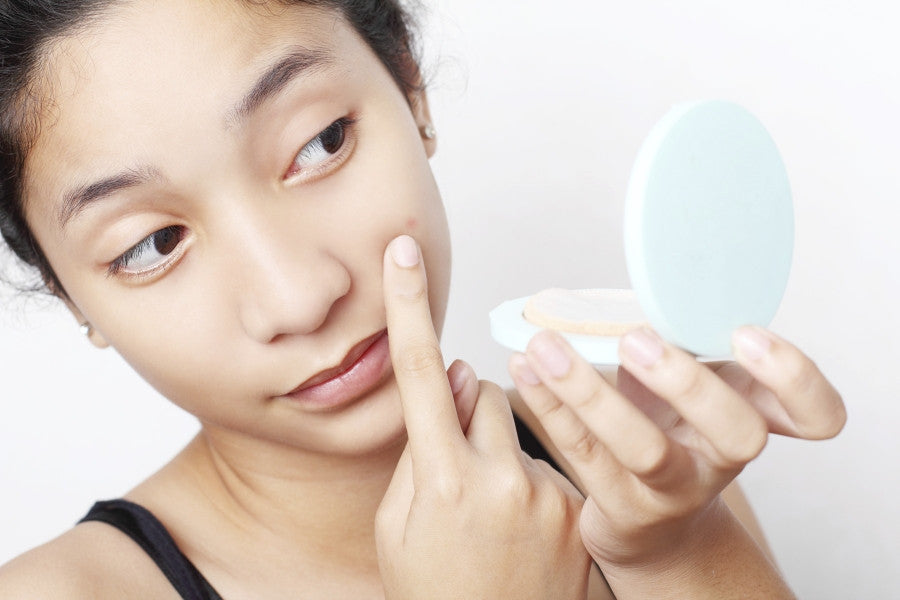 Brush On Block image of woman studying cheek blemish in hand mirror.