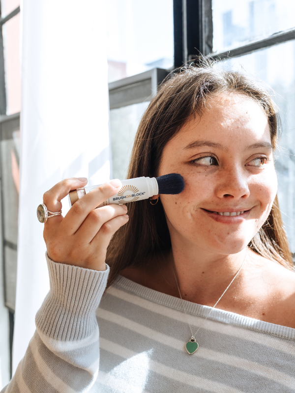 A woman applying Brush On Block SPF 50 mineral powder sunscreen to her cheek 