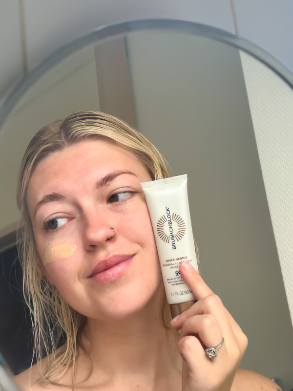 Woman holding a tube of Brush On Block Sheer Genius SPF 50 mineral moisturizing sunscreen lotion by her face 