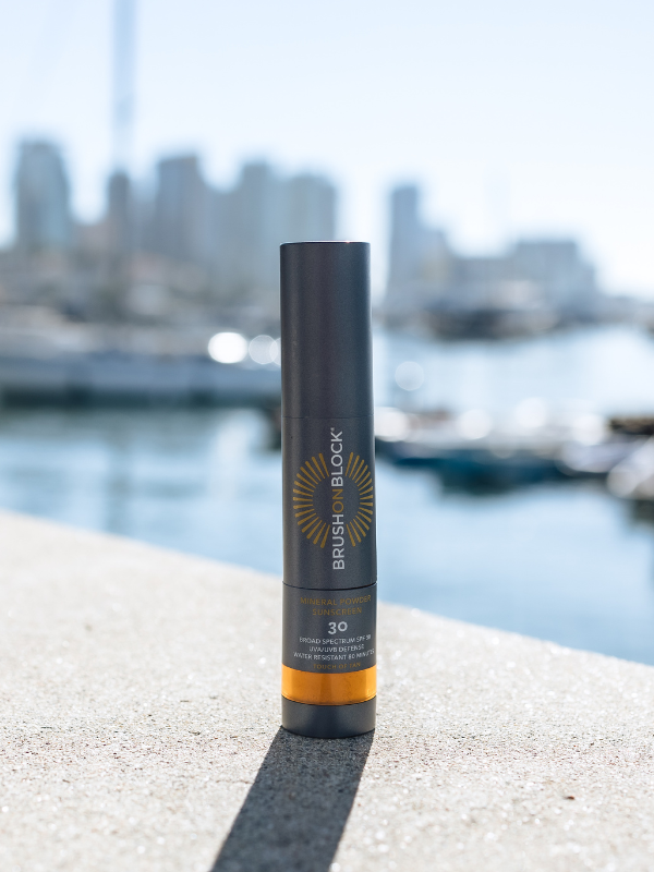 Brush On Block SPF 30 Mineral Powder Sunscreen sitting on a sea wall ledge with boats in the background 