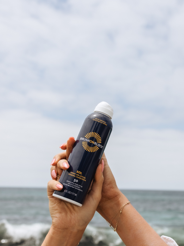 Hands holding Brush On Block SOL SPF 50 Spray-On Lotion in the air over the surf. 