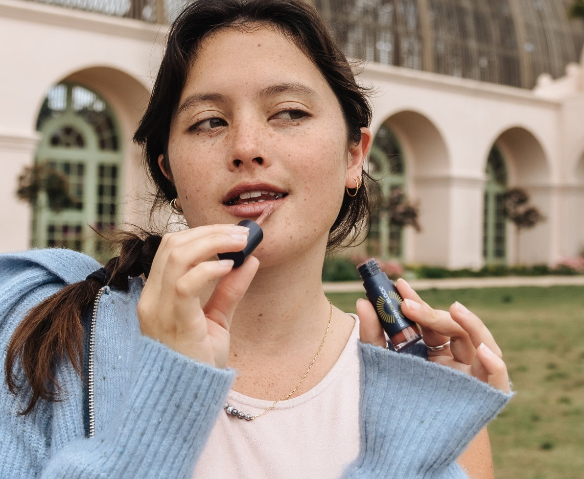 Woman applying Sun Shine SPF 30 lip oil outdoors with a building in the background